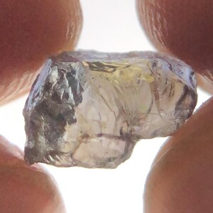 May include: A raw, uncut gemstone with a translucent, multifaceted appearance. The stone displays a pale, light-toned coloration with subtle hints of yellow and purple throughout its irregular, crystalline structure and sharp, jagged edges.