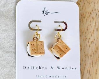May include: A pair of s'mores dangle earrings featuring miniature graham cracker, marshmallow, and chocolate layers. The earrings are attached to a white display card printed with the text 'dw', 'Delights & Wonder', and 'Handmade in Colorado'.