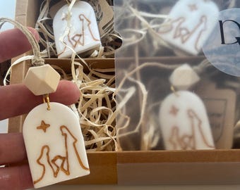 May include: White ceramic ornament with a gold outline of a nativity scene, a star, and a wooden bead. The ornament is hanging from a natural twine cord. The ornament is in a brown box with other ornaments.
