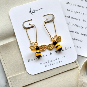 May include: A pair of gold-toned safety pin earrings featuring bee charms with yellow and black striped bodies and clear rhinestone-encrusted wings. The earrings are displayed on a white card printed with the text Delights & Wonder Handmade in Colorado.