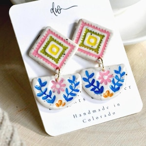 May include: A pair of dangle earrings on a white display card labeled "dw" and "Handmade in Colorado". Each earring features a white diamond-shaped top with pink, green, and yellow geometric patterns, connected to a white semi-circle with blue floral designs.