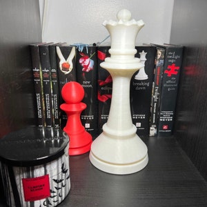 Könnte beinhalten: Eine weiße Schachkönigin und ein roter Bauer stehen auf einem Regal vor einer Reihe von Büchern. Eine schwarze Kerze mit Fledermausdesign und dem Text "Vampire Blood" ist ebenfalls vorhanden. Die Bücher gehören zur Twilight-Reihe.