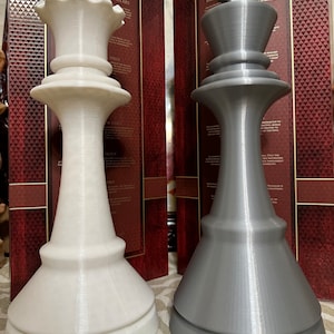 Könnte beinhalten: Eine weiße Schachkönigin und ein grauer Schachkönig werden nebeneinander dargestellt. Die Königin hat eine kronenartige Spitze, während der König eine kreuzförmige Spitze hat. Beide Figuren haben ein detailliertes, strukturiertes Design. Der Hintergrund enthält Text auf einer dunkelroten Oberfläche.