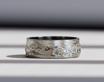 May include: A silver ring with a mountain range design. The ring is wide and has a textured surface.
