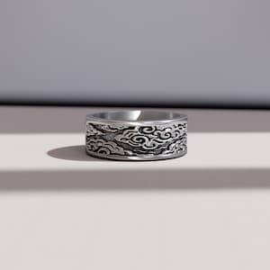 Può includere: Un anello d'argento con un motivo a nuvole dettagliato che avvolge la fascia. L'anello ha una finitura lucida e un design a fascia larga. Il motivo a nuvole è in una tonalità più scura, creando contrasto con l'argento.
