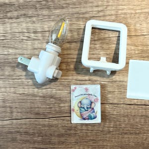 May include: A white night light with a clear bulb, a white frame, and a small rectangular piece. A small rectangular piece has an illustration of a sleeping elephant on a crescent moon with the text "Sweet Dreams Little One".