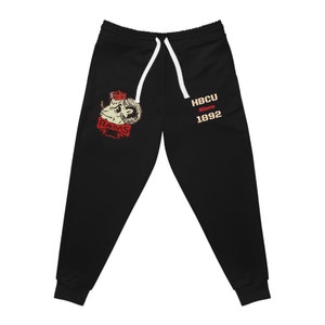 Può includere: Pantaloni della tuta neri con una grafica rossa e bianca di un ariete con una corona sulla testa e il testo "RAMS" e "HBCU Since 1892".