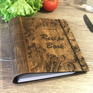 Puede incluir: Un libro de recetas de madera con una cubierta marrón y una banda elástica negra. La cubierta está decorada con un diseño grabado de verduras y el texto "The Bilan Recipe Book".