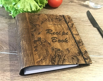 May include: A wooden recipe book with a brown cover and a black elastic band. The cover is decorated with an engraved design of vegetables and the text "The Bilan Recipe Book".