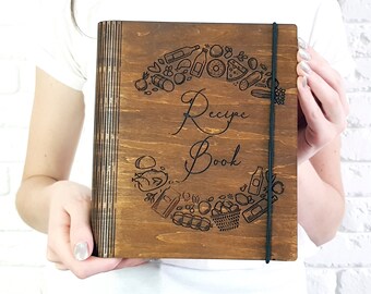 May include: A wooden recipe book with a dark brown finish. The cover features the words "Recipe Book" in elegant script, surrounded by engraved food illustrations. A black elastic band secures the book.