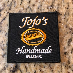 May include: Black fabric patch with white embroidery that reads "Jojo's Handmade Music". A gold and white circular design with a stylized musical instrument is in the center.