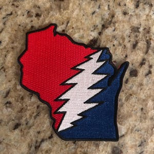 May include: Embroidered patch in the shape of Wisconsin, divided into red and blue sections with a white lightning bolt design. The patch has a black border and is likely for clothing or accessories.