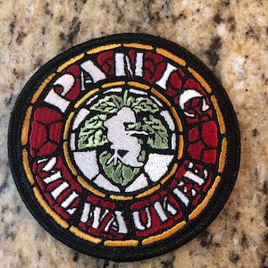 May include: A round, black, embroidered patch with a stained glass design. The center features a green leaf with a white dove inside. The words "Panic! At The Disco" are written in a circle around the leaf.