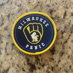 May include: A blue and yellow embroidered patch with the words "Milwaukee" and "Panic" and a white baseball glove graphic.