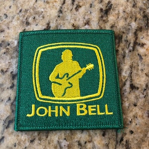 May include: Green and yellow embroidered patch with a silhouette of a person playing a guitar inside a square. The text "JOHN BELL" is below the square.