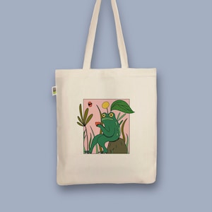 Könnte beinhalten: Beige Tragetasche mit einem Cartoon-Frosch-Design. Der grüne Frosch wird mit einem Getränk dargestellt, umgeben von Pflanzen, innerhalb eines rosa Rechtecks. Die Tasche hat lange Griffe, ideal zum Tragen von Gegenständen.