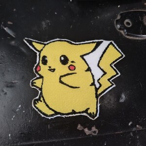 May include: A yellow embroidered patch featuring a cartoon character with pointed ears, a lightning bolt-shaped tail, and red circular cheeks. The design is outlined in black thread and includes black eyes and a small mouth on a textured fabric surface.