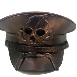 Puede incluir: Un sombrero de estilo militar de color bronce con un diseño de calavera y huesos cruzados en la parte delantera. El sombrero tiene un ala ancha y una corona alta.