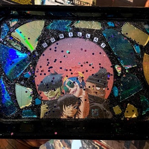 Puede incluir: Una bandeja de metal negro con una superficie de resina brillante. La bandeja presenta una imagen circular de la banda Gorillaz con el nombre de la banda escrito en letras blancas. La bandeja est&aacute; decorada con trozos de vidrio iridiscente rotos.