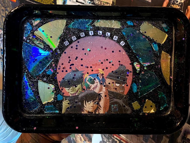 Puede incluir: Una bandeja de metal negro con una superficie de resina brillante. La bandeja presenta una imagen circular de la banda Gorillaz con el nombre de la banda escrito en letras blancas. La bandeja est&aacute; decorada con trozos de vidrio iridiscente rotos.