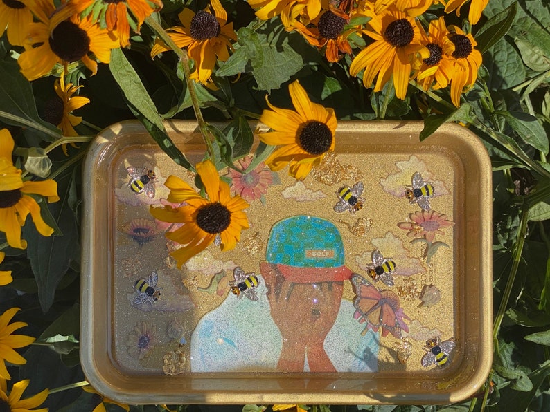 Puede incluir: Bandeja dorada con una superficie brillante que presenta una ilustraci&oacute;n de dibujos animados de una persona que lleva un sombrero con estampado azul y verde. La bandeja est&aacute; decorada con abejas, nubes y flores.