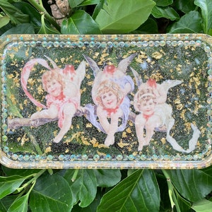 Puede incluir: Una bandeja rectangular con un borde de purpurina dorada y un interior de resina transparente. La bandeja presenta tres figuras de querubines con alas, cada una con una pose y una expresi&oacute;n diferentes. Los querubines est&aacute;n rodeados de copos de pan de oro.