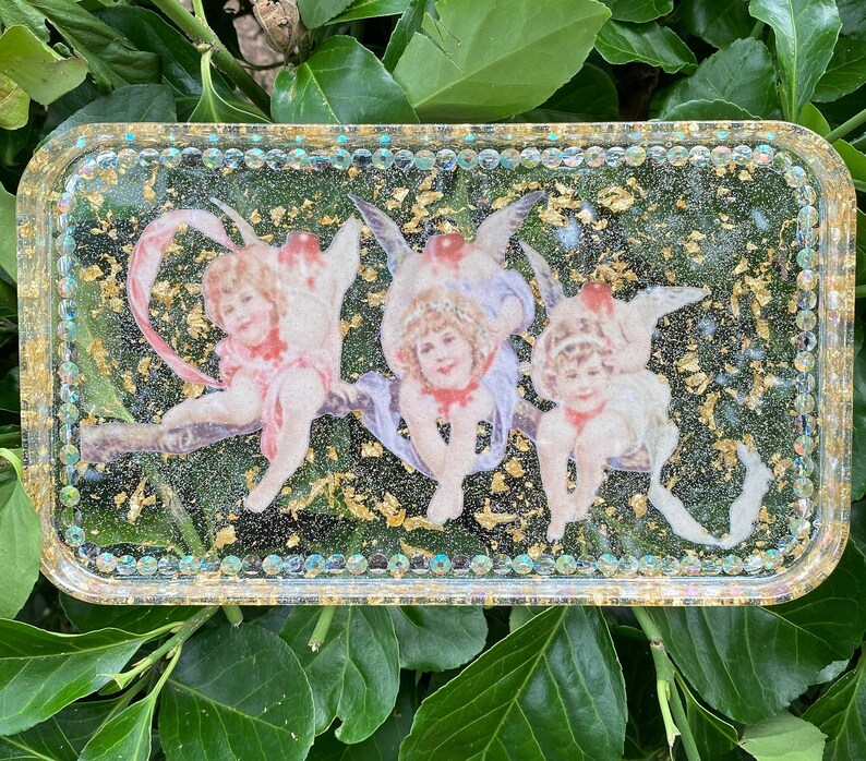 Puede incluir: Una bandeja rectangular con un borde de purpurina dorada y un interior de resina transparente. La bandeja presenta tres figuras de querubines con alas, cada una con una pose y una expresi&oacute;n diferentes. Los querubines est&aacute;n rodeados de copos de pan de oro.