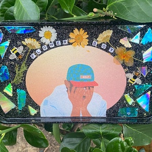Puede incluir: Una bandeja negra con una superficie brillante y un dise&ntilde;o colorido. El dise&ntilde;o presenta a una persona con un sombrero de patr&oacute;n azul y blanco, un fondo rosa y naranja, y las palabras "FLOWER BOY".