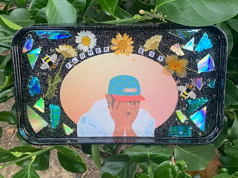 Puede incluir: Una bandeja negra con una superficie brillante y un dise&ntilde;o colorido. El dise&ntilde;o presenta a una persona con un sombrero de patr&oacute;n azul y blanco, un fondo rosa y naranja, y las palabras "FLOWER BOY".