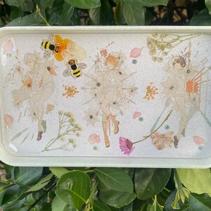 Puede incluir: Una bandeja rectangular blanca con una superficie brillante. La bandeja presenta un dise&ntilde;o de flores prensadas con tres figuras y dos abejas. Las figuras est&aacute;n rodeadas de flores y hojas secas.