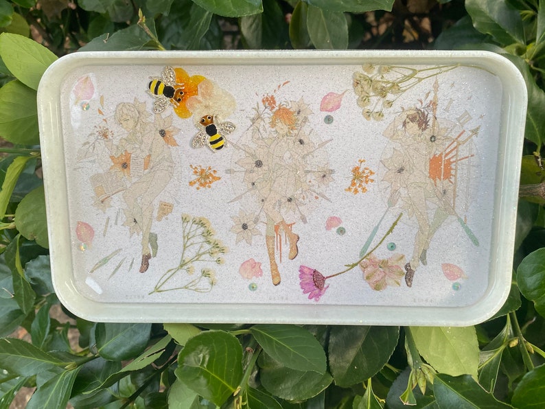 Puede incluir: Una bandeja rectangular blanca con una superficie brillante. La bandeja presenta un dise&ntilde;o de flores prensadas con tres figuras y dos abejas. Las figuras est&aacute;n rodeadas de flores y hojas secas.