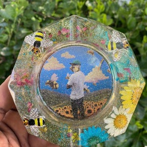 May include: Hexagonal ashtray with a resin finish, featuring a central image of a person in a field with bees and a blue sky. The ashtray is decorated with glitter, flowers, and three bee embellishments.