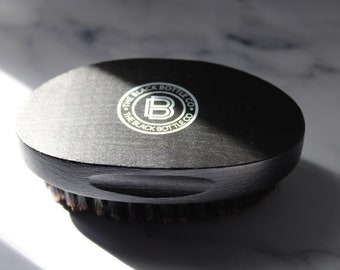 May include: A black oval-shaped brush with a wooden handle and black bristles. The handle has a white logo that reads "The Black Bottle Co."