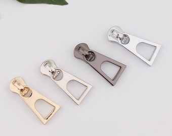 May include: Four zipper pulls in different finishes: gold, silver, black, and antique brass. Each pull has a rectangular shape with a rounded top and a small hole at the top.