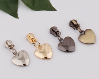 May include: Four heart-shaped zipper pulls in different finishes: gold, silver, antique brass, and black. Each pull has a small chain attached.