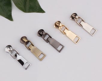 Könnte beinhalten: Fünf Reißverschluss-Zipper in verschiedenen Ausführungen: Silber, Bronze, Schwarz, Gold und Weißgold. Die Zipper sind alle gleich groß und haben die gleiche Form, mit einem rechteckigen Schieber und einer kleinen Schlaufe oben.