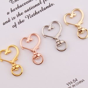 Könnte beinhalten: Vier herzförmige Metallverschlüsse in Gold, Silber und Roségold. Die Verschlüsse sind mit einer herzförmigen Schlaufe und einem Federverschluss ausgestattet.