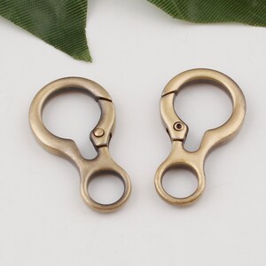 May include: Two antique gold-toned metal swivel clasps with a round loop on each side. The clasps are open and ready to be attached to a bag, keychain, or other item.