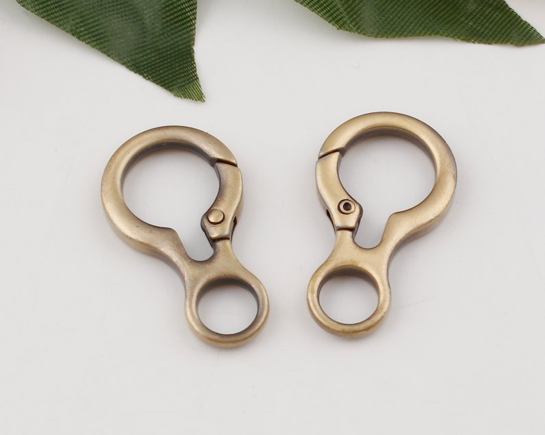 May include: Two antique gold-toned metal swivel clasps with a round loop on each side. The clasps are open and ready to be attached to a bag, keychain, or other item.