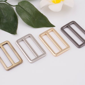May include: Four metal slider buckles in different finishes: antique brass, silver, gold, and gunmetal. Each buckle is rectangular with two parallel slots.