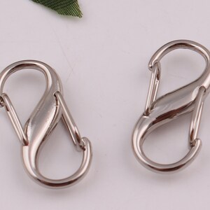 Pu&ograve; includere: Due chiusure metalliche a forma di S color argento, ciascuna con una finitura liscia e arrotondata. Queste chiusure sono comunemente utilizzate per attaccare oggetti a portachiavi, borse o altri accessori.