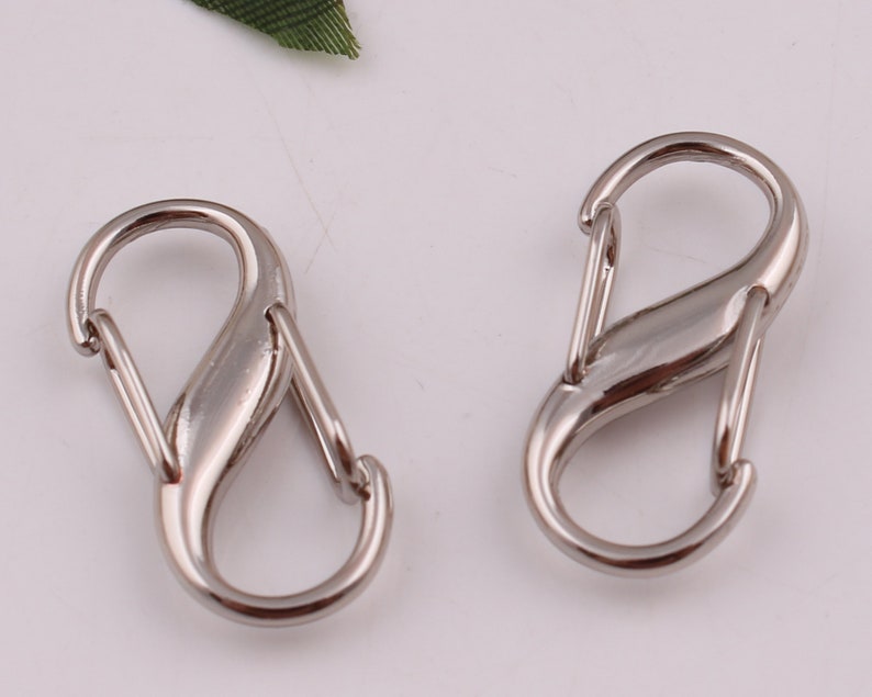 Pu&ograve; includere: Due chiusure metalliche a forma di S color argento, ciascuna con una finitura liscia e arrotondata. Queste chiusure sono comunemente utilizzate per attaccare oggetti a portachiavi, borse o altri accessori.