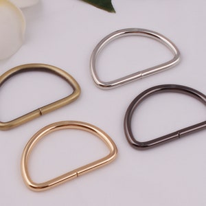 May include: Four metal D-rings in different finishes: silver, gold, antique brass, and black. These rings are commonly used for attaching straps, chains, or other hardware to bags, purses, or other accessories.