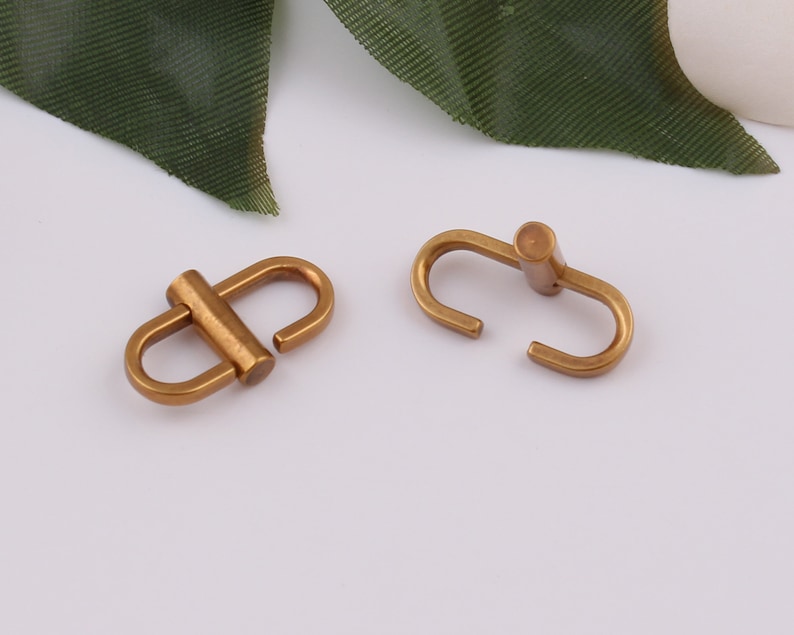 Op de afbeelding: Twee goudkleurige metalen gespen, elk met een afgeronde, cilindrische stang in het midden. De gespen zijn ontworpen om twee stukken materiaal aan elkaar te verbinden.