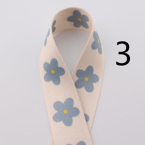 Op de afbeelding: Een wit katoenen lint met een blauw bloemenpatroon. Het lint heeft een gele kern in elke bloem. Het nummer 3 is zichtbaar in de afbeelding.