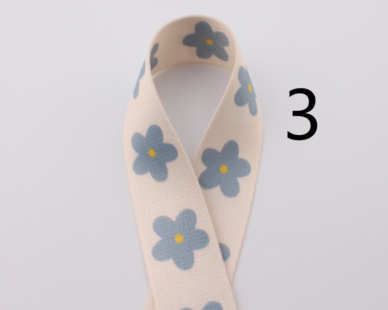 Op de afbeelding: Een wit katoenen lint met een blauw bloemenpatroon. Het lint heeft een gele kern in elke bloem. Het nummer 3 is zichtbaar in de afbeelding.