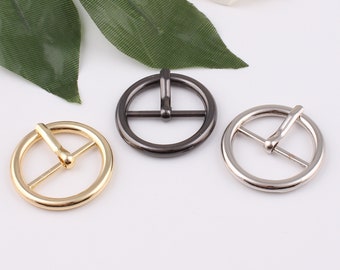 May include: Three metal belt buckles in different finishes: gold, silver, and black. Each buckle has a round shape with a center bar.