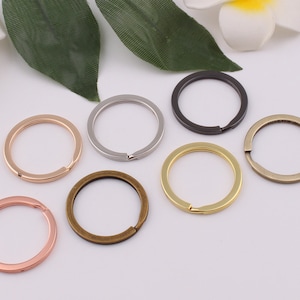 Puede incluir: Siete llaveros de diferentes colores: plata, oro, oro rosa, bronce y negro. Los llaveros son todos del mismo tamaño y forma, y están dispuestos en círculo sobre un fondo blanco.