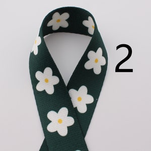 Op de afbeelding: Groen lint met een witte madeliefjesprint. Het lint heeft een gele kern op elke madeliefje. Het nummer 2 staat in zwart op een witte achtergrond rechts van het lint.