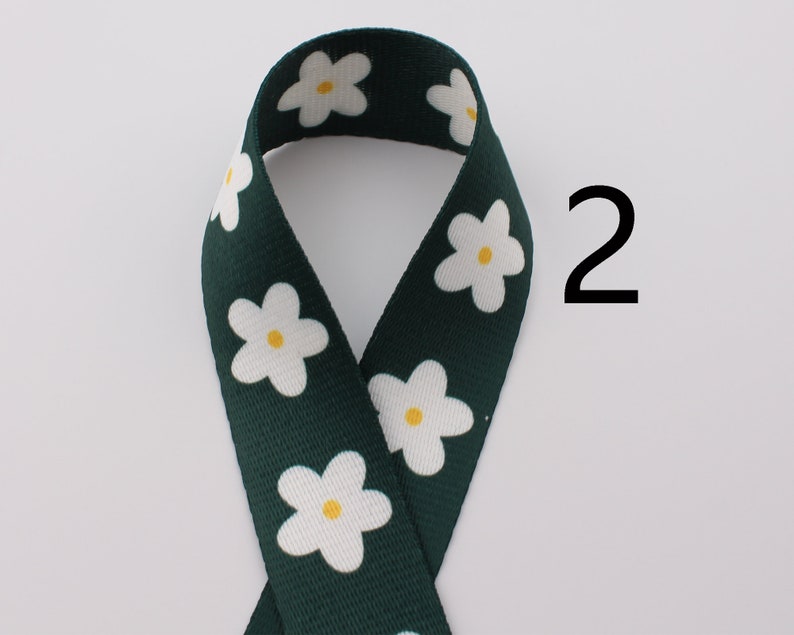 Op de afbeelding: Groen lint met een witte madeliefjesprint. Het lint heeft een gele kern op elke madeliefje. Het nummer 2 staat in zwart op een witte achtergrond rechts van het lint.